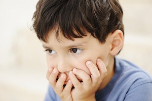 Lille infantil autistisk dreng med mørkt hår og øjne, sidder med hænderne foran munden og ser trist ud. 