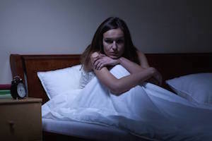 Kvinde sidder oprejst i sin seng midt om natten med dynene over benene, fordi hun ikke kan sove 