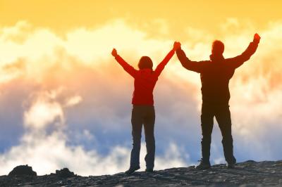 Mand og dame står på toppen af et bjerg hånd i hånd og med armene over hovedet, mens de kigger på en farverig og skydækket himmel 